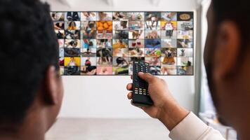 A man sits in front of a television screen that is displaying a grid of smaller screens, each showing a different image or . He is using a remote control to change the channel. photo