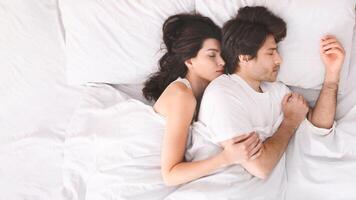 A couple is sleeping peacefully in bed together, with the woman nestled against the mans back, wearing white shirts and are completely relaxed, suggesting a comfortable and loving relationship. photo