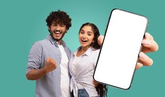 A young Indian couple stand against a teal background. They both look directly at the camera and smile. The woman holds a smartphone in front of them, displaying a blank white screen photo