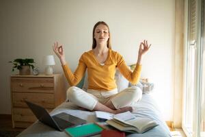 A teen girl is sitting on her bed, meditating with closed eyes and a serene expression. She is surrounded by open books, a laptop, and notebooks, indicating she is taking a break from studying photo
