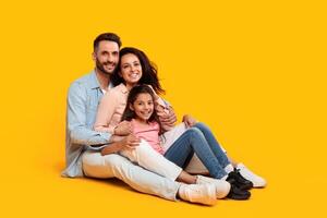 A family of three, a father, a mother, and their daughter, are sitting together on a bright yellow background. They are all smiling and looking at the camera. The father has his arm around the mother photo