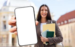 A young woman smiles broadly while holding a blank smartphone in one hand and several colorful files in the other, standing in front of an attractive modern building on a sunny day. photo