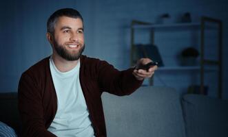 A bearded man sits on a couch, smiling while pointing a remote control towards a television in a cozy living room at night. photo