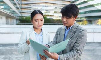 Two Asian business professionals, wearing formal attire, are standing in front of a modern office building. They are attentively reviewing a folder, possibly discussing important documents photo