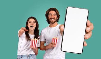 A smiling couple enjoying a movie on a smartphone. They are both holding red and white striped containers of popcorn, and the man is holding the phone forward for the viewer to see photo