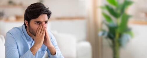A young Indian man sits on a couch, deep in thought and contemplation. His expression shows concern and introspection, with a green plant softly blurred in the background for a calming effect. photo