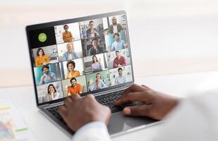 Entrepreneur is sitting at a desk using a laptop to participate in a conference. The laptop screen displays multiple participants in a grid layout, each in their own frame. photo