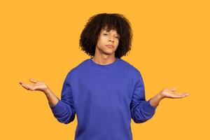 A young man with curly hair, wearing a blue sweatshirt, stands against a bright yellow backdrop. He is gesturing with both hands, palms facing upward, as if shrugging or expressing uncertainty photo