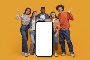 Five happy young friends stand in front of a vibrant orange background, holding a large smartphone with a blank white screen. They are smiling and laughing photo