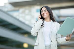 A confident woman in a light suit is engaged in a phone conversation, smiling while holding folders outside a modern building. photo