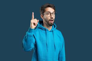 A young man wearing a blue hoodie is pointing upward with his right index finger while looking surprised or excited. His glasses add an intellectual touch to his expression, which conveys enthusiasm photo