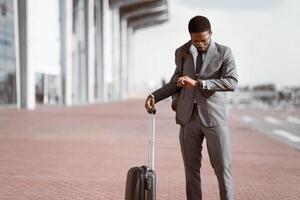 Black businessman checking time on his watch with rolling suitcase outside a modern building, preparing for a business trip. photo