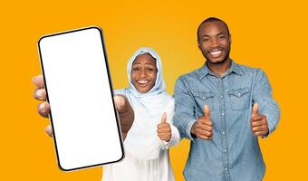 Black woman wearing a headscarf and a man, are holding a smartphone with a blank screen and giving thumbs up. They are smiling and looking directly at the camera photo