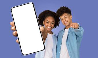 A young black woman and man are standing side-by-side, smiling and pointing at the camera. They are holding a smartphone with a blank screen in front of them. The background is a light blue color. photo