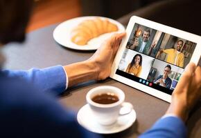 Black man sits at a table in a casual setting, participating in a conference on a tablet. The screen displays four participants, each in a different frame. photo