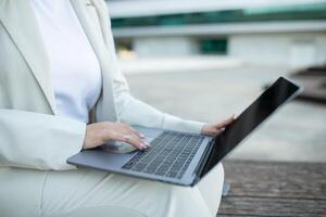 A woman wearing a white suit is sitting on a bench outside. She is using a laptop and appears to be working or browsing the internet. The background is blurred, suggesting a modern urban environment. photo