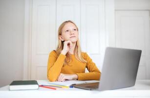 A teen girl sits at a desk with a laptop and a notebook. She has one hand on her chin and is looking upward, lost in thought. The desk is also adorned with colored pencils and a notebook. photo
