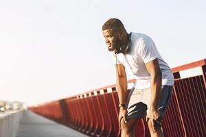 A man in athletic wear rests on a bridge railing after a run. He is wearing headphones and has a serious expression on his face. photo