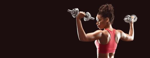 Rear view of african sporty girl working out with dumbbells over black studio background photo