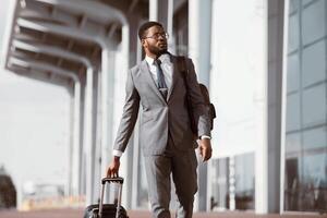 African American businessman walking with rolling suitcase outside a modern building, ready for a business trip or travel. photo