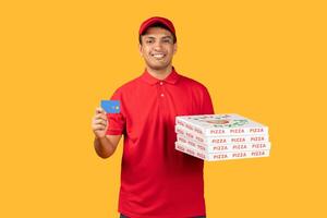 Delivery man standing and holding a pizza box in one hand and a card in the other. He is dressed in red unform, yellow background photo