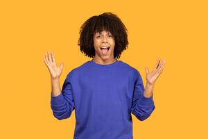 Young man with curly hair wearing a blue sweatshirt and standing in front of a yellow background. He has a surprised expression on his face and is holding both of his hands up in the air. photo