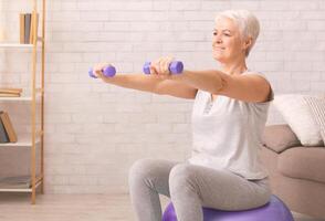 Senior woman in athletic attire is performing exercises on an exercise ball with dumbbells in living room at home. She balances carefully while engaging her core muscles and limbs photo