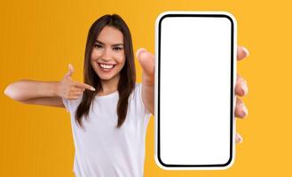Check This Website. Portrait of smiling female holding smartphone with white blank screen in hand, showing device close up to camera and pointing finger at gadget with empty free space for mock up photo