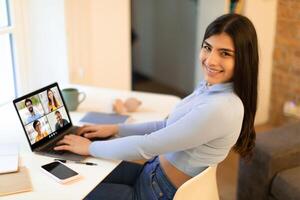 Smiling woman sits at a desk in a home office, smiling and looking at the camera while participating in a conference on her laptop. The conference window shows several faces photo