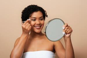 A young Hispanic woman is standing in front of a mirror, carefully applying makeup to her face. She is focused and precise, using various tools and products to enhance her features. photo