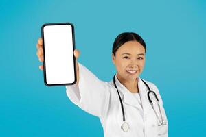 Asian woman doctor wearing a white coat and stethoscope smiles at the camera while holding a smartphone with a blank screen in her right hand. The background is a bright blue. photo