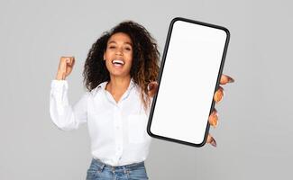 Black woman stands holding a cell phone with a blank screen in her hand, raising it towards eye level. The phone is a modern smartphone with a sleek design, displaying no content on the screen. photo