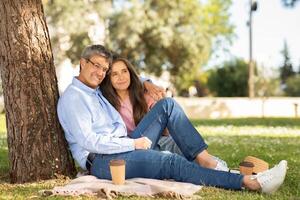 A couple sits together on a blanket, leaning against a tree in a lush park. They share a warm moment, smiling while sipping coffee, enjoying the sunny day. photo