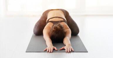 A lady is engaged in a relaxing yoga session, performing the Childs Pose on a dark yoga mat in a brightly lit studio, suggesting a tranquil and centered exercise routine. photo