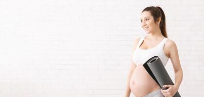 A pregnant woman is standing while holding a yoga mat in her hands. She appears focused and prepared for a yoga session, showing dedication to her practice even during pregnancy, copy space photo