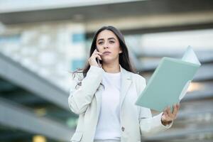 A woman in a light suit engages in a phone conversation while holding a file, standing outside a modern building. photo