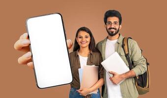 Two smiling Indian students stand in front of a brown background. They are holding books and a smartphone with a blank screen photo