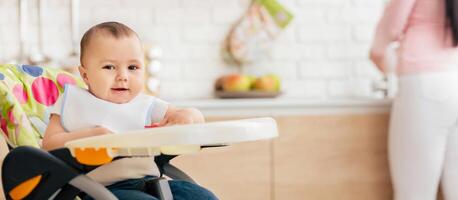 A baby boy is enjoying his meal in a child chair in the kitchen. The setting is warm and inviting, capturing a moment of contentment and family routine. photo