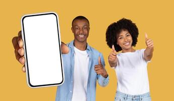 Smiling black couple standing in front of a yellow background. The man is holding a smartphone with a blank screen, while the woman is giving a thumbs-up with both hands photo