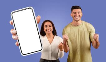 A young woman holds a smartphone with a white screen in front of her while smiling and celebrating with a man. The man has his fists clenched in the air and is also smiling. photo