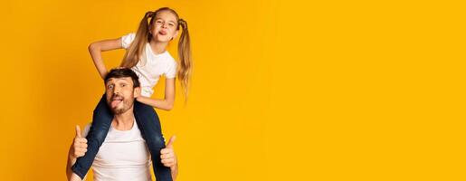 A joyful father gives his daughter a piggyback ride, both making silly faces and showing their thumbs up. The vibrant yellow backdrop adds to the cheerful atmosphere of their playful interaction. photo
