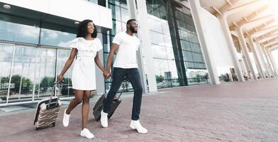 Black young couple smiling, walk towards the camera with their luggage in hand. They are outside of an airport building, with large glass windows and doors visible behind them, copy space photo