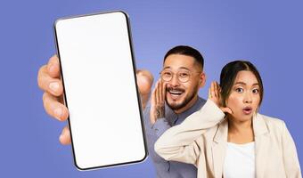 Asian man with a beard and glasses shouts into the screen of a smartphone held by an unseen hand. A woman with her hand to her ear listens intently, with a surprised expression on her face. photo