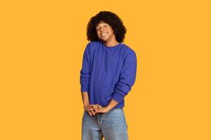 This image shows a young man smiling in front of a yellow background. He is wearing a blue sweatshirt and jeans. He has a happy and relaxed expression, and appears to be confident and at ease. photo