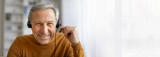 A senior man with gray hair is wearing a headset and smiling at the camera. He is likely working from home, as he is sitting in front of a window, copy space photo