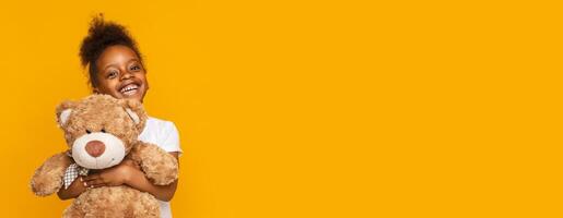 A cheerful young black girl is happily hugging a soft teddy bear while standing in front of a vibrant yellow background. Her bright smile conveys a sense of joy and innocence, copy space photo