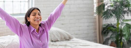 Middle eastern woman raises her arms in a joyful stretch while sitting on her bed, basking in the soft morning light streaming through the window, creating a refreshing and uplifting atmosphere. photo