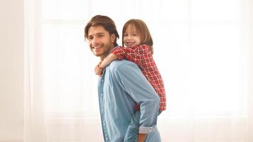 Young father riding his little daughter on his back at home, staying next to window and smiling, copy space photo