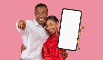 A happy black couple is standing in front of a pink background. The man is pointing towards the camera with his left hand. The woman is holding up a smartphone with a blank screen photo