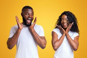 A man and a woman are standing in front of a yellow background, both looking shocked and with their hands on their cheeks. photo